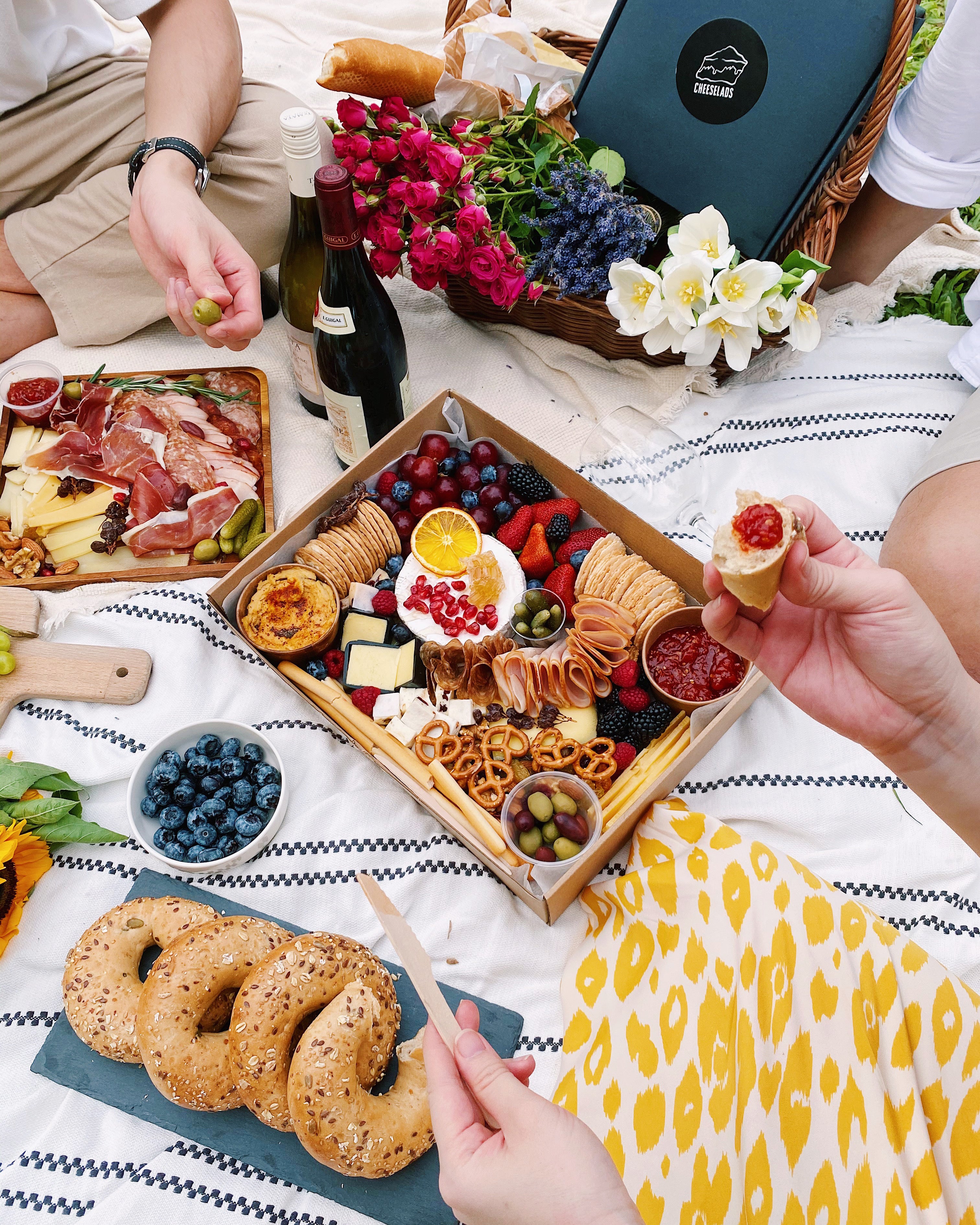 A person holding a small bite of food at a picnic with a large box of charcuterie.
