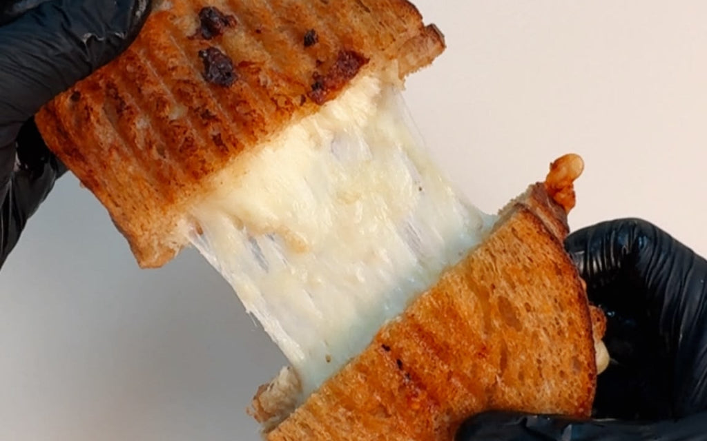 Hands in black gloves pulling apart a toasted grilled cheese sandwich, stretching thick, melted white cheese between the two golden-brown halves.
