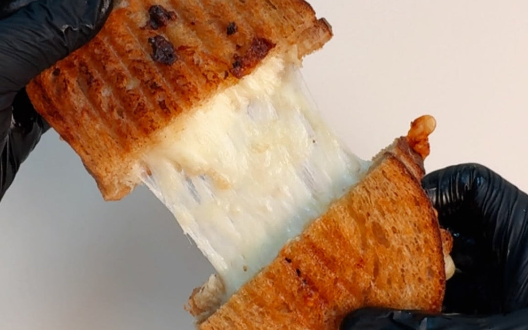 Hands in black gloves pulling apart a toasted grilled cheese sandwich, stretching thick, melted white cheese between the two golden-brown halves.